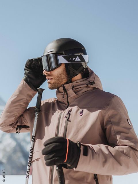 Gros plan sur une personne portant un casque de ski gris, des lunettes de ski réfléchissantes et une veste foncée, avec un paysage enneigé flou en arrière-plan.