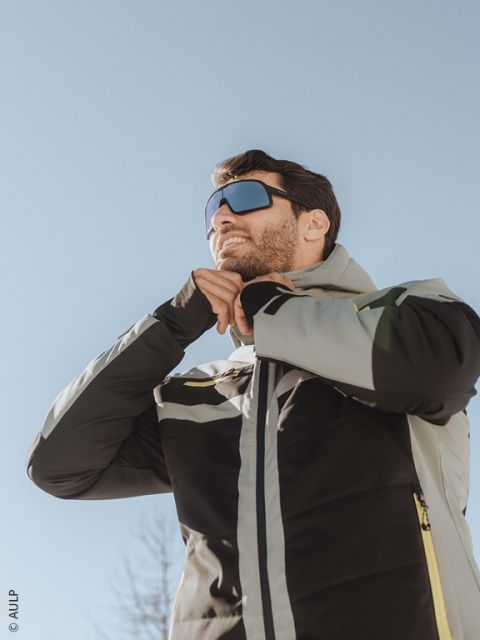 Un skieur portant un casque blanc, des lunettes réfléchissantes, une veste et des gants noir et marron ferme sa veste à l'extérieur avec des montagnes enneigées en arrière-plan.