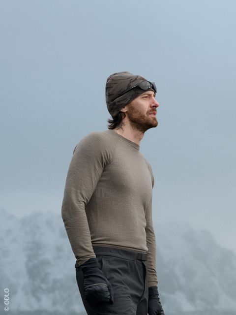 Un homme et une femme en vêtements d'hiver décontractés sont assis à l'extérieur sur un mur de pierre, souriant et discutant. Une paire de skis est appuyée contre le mur et des montagnes enneigées sont visibles en arrière-plan.
