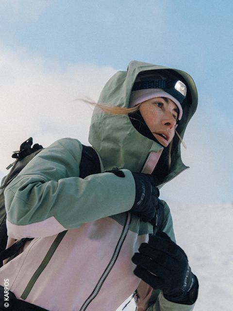 Une femme portant une veste Dynafit bouffante, des lunettes de soleil et des gants se prépare pour une randonnée dans un paysage montagneux enneigé, avec des sommets enneigés en arrière-plan. Elle porte un sac à dos et des bâtons de ski.