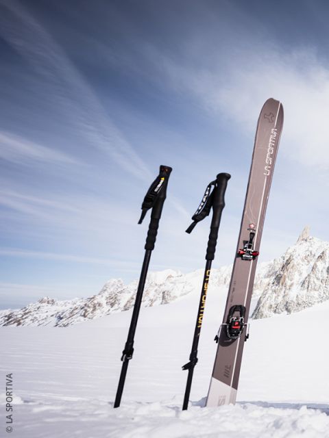 Deux personnes en vêtements de ski colorés et avec des sacs à dos montent une montagne enneigée en s'aidant de bâtons. En arrière-plan, on aperçoit un grand sommet montagneux escarpé.