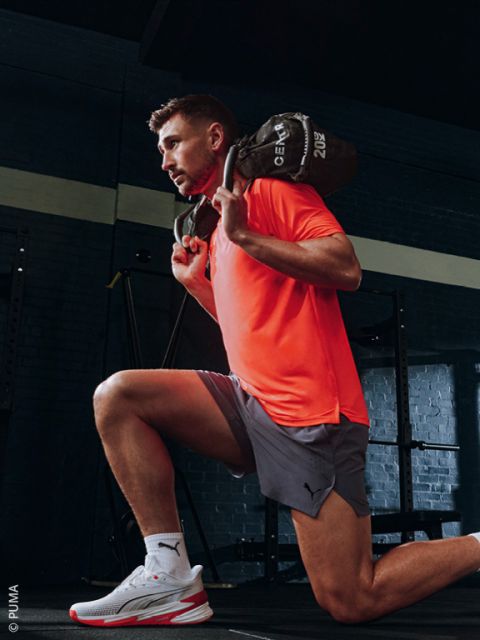 Un homme en T-shirt orange et short gris fait une fente dans une salle de sport, tenant un sac rempli de poids sur ses épaules.