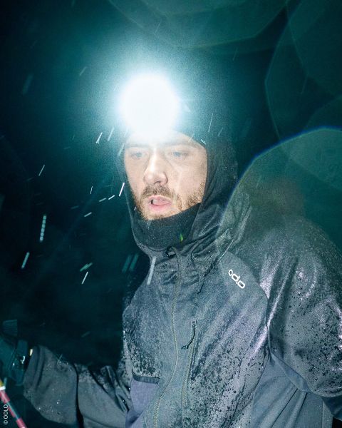 Une femme portant une lampe frontale, un bandeau à motifs et un gilet de trail running lève les yeux vers le ciel nuageux.