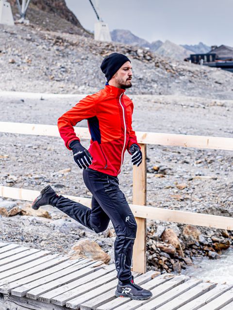 Un homme en veste rouge, pantalon noir, gants et bonnet marche sur un pont en bois dans une région rocheuse et montagneuse.