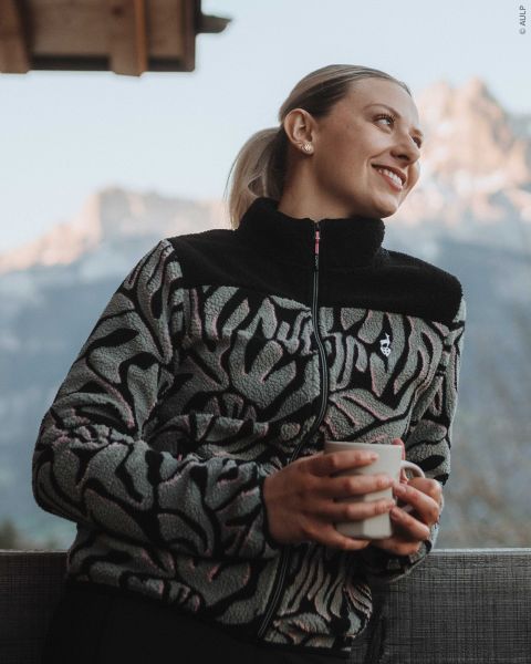 Une femme portant une veste polaire zippée à motifs tient une tasse et regarde les montagnes en arrière-plan.